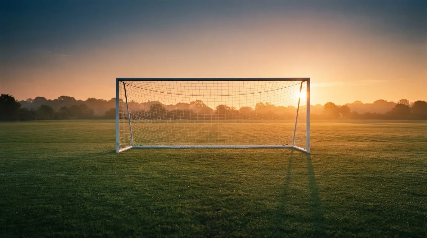 Ein Fußballtor mit Netz bei Sonnenuntergang auf einem leeren Platz, die Schatten der Torpfosten liegen lang auf dem Gras.