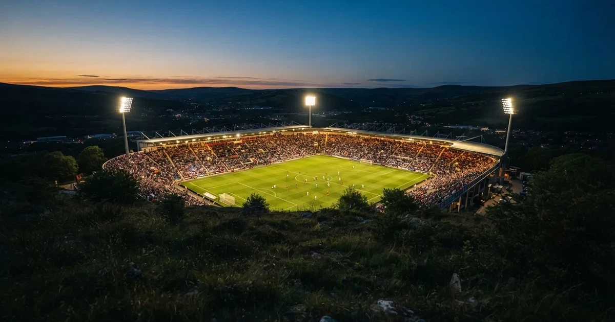 Ein Fußballstadion unter Flutlicht mit hellen Rängen, von einem Hügel aus gesehen, darüber ein dunkelblauer Abendhimmel.