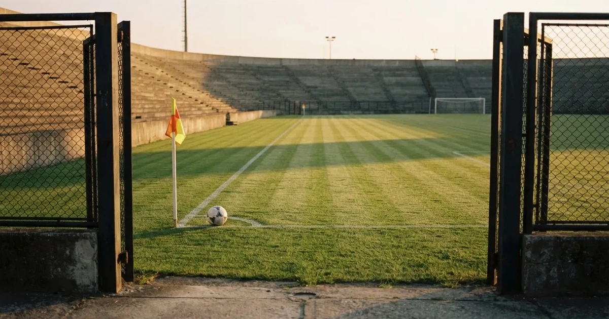 Blick durch ein Stadiontor auf einen grünen Rasen im Abendlicht, ein Ball ruht an der Eckfahne, die Tribünen sind leer.