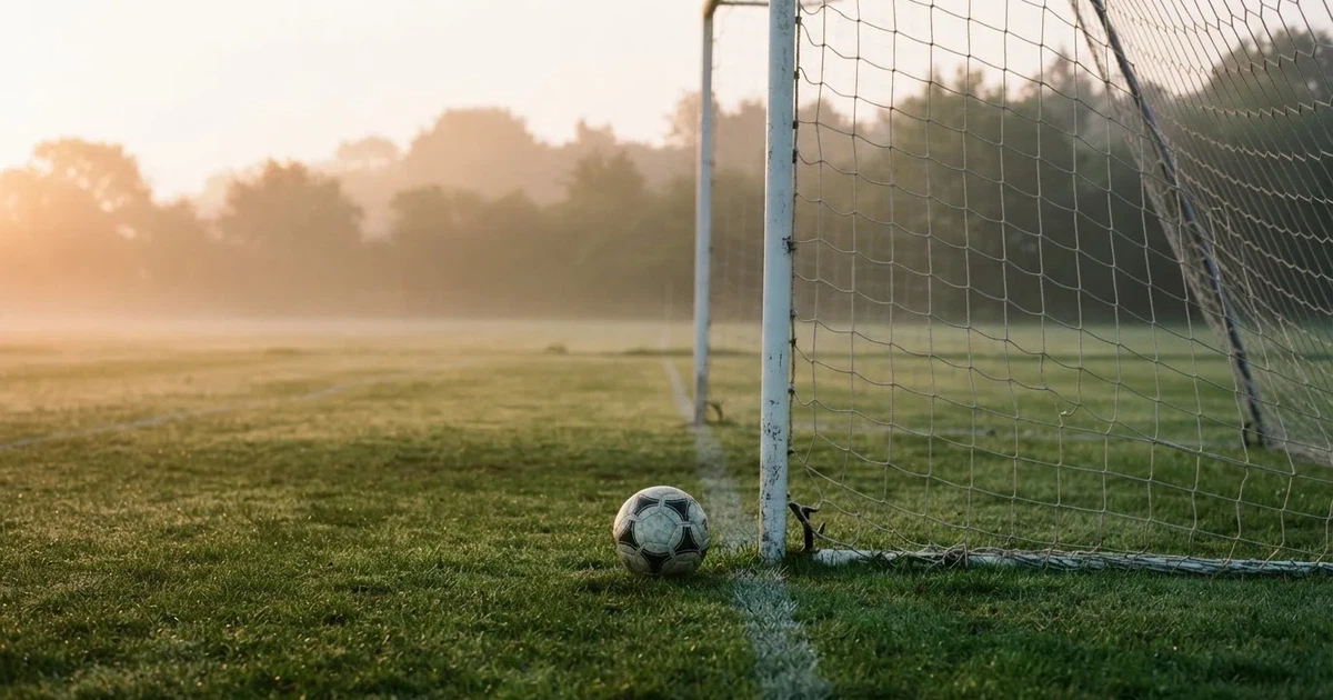 Ein Fußballtor mit gespanntem Netz steht auf einem leeren Rasenplatz, davor ein einzelner Ball, Morgendunst liegt über dem Feld.