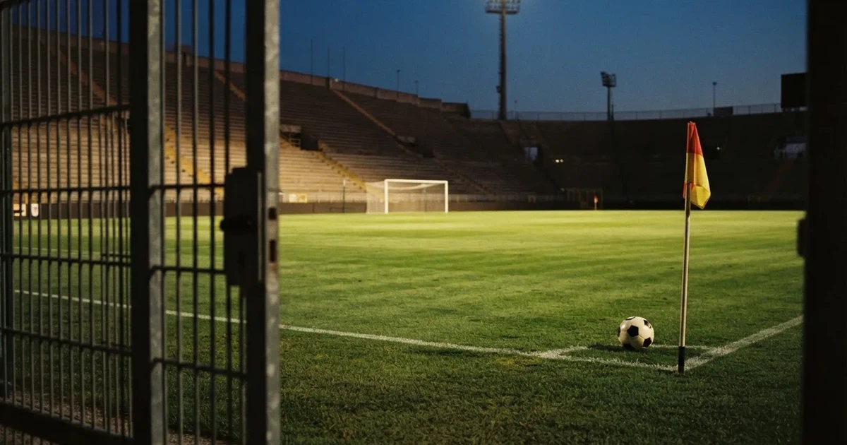Blick durch ein Stadiontor auf einen grünen Rasen im Abendlicht, ein Ball liegt an der Eckfahne, die Ränge sind leer.