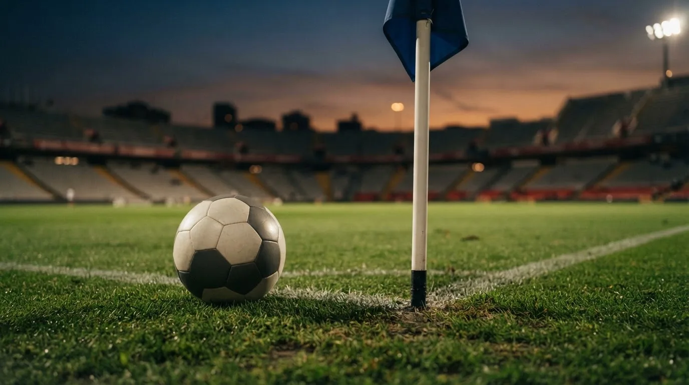 Ein Fußball liegt neben einem Eckfahnen-Pfosten auf einem Rasenplatz, im Hintergrund ein leeres Stadion in der Dämmerung.