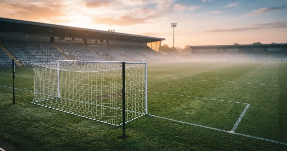 Ein Fußballplatz mit gepflegtem Rasen und einem Tor, über dem Spielfeld liegt Morgennebel, im Hintergrund eine leere Tribüne.