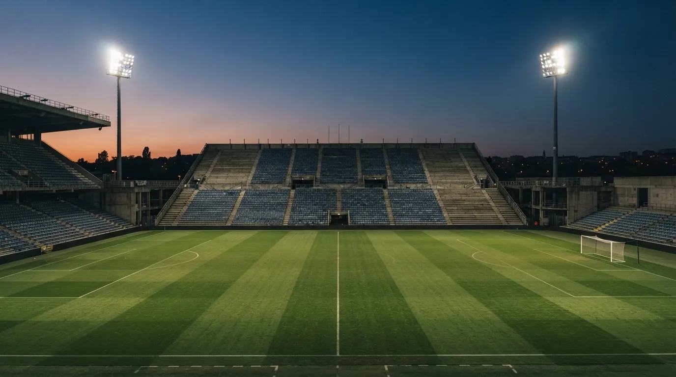 Eine leere Stadiontribüne unter Flutlicht aus der Ferne betrachtet, die Rasenfläche leuchtet grün in der Dämmerung.
