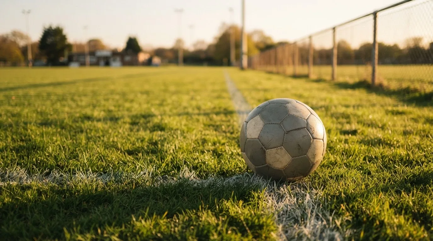 Ein Fußball liegt am Rand eines Rasens neben einer weißen Linie, im Hintergrund ein Zaun in der Abendsonne.