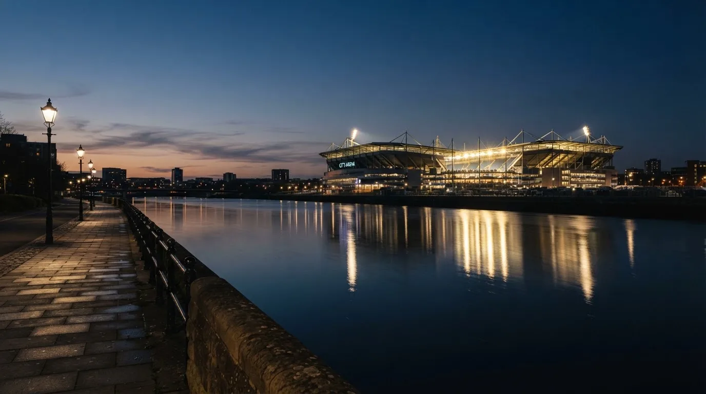 Ein Stadion unter Flutlicht aus der Ferne gesehen, im Vordergrund ein ruhiger Fluss und ein Fußweg.