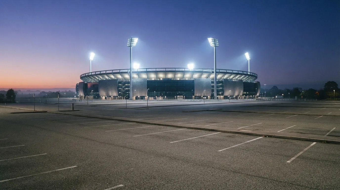 Ein Stadion unter Flutlicht aus der Ferne gesehen, davor ein leerer Parkplatz in der Dämmerung.