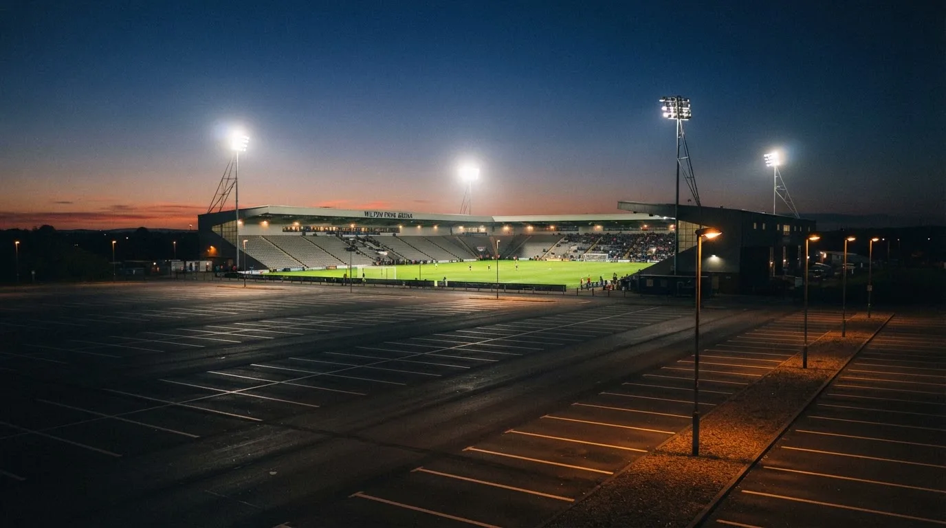 Ein Fußballstadion unter Flutlicht, aus großer Entfernung bei Einbruch der Dunkelheit gesehen, davor ein leerer Parkplatz.
