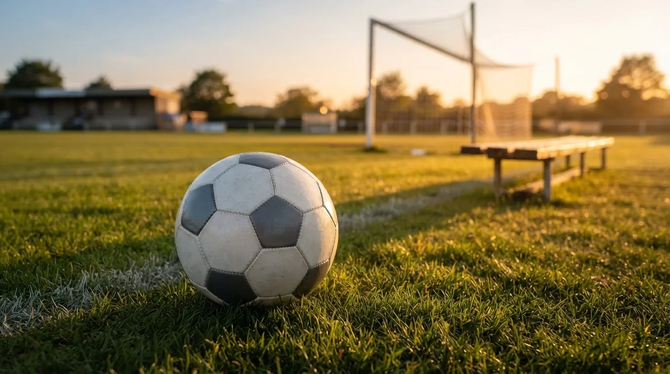 Ein Fußball liegt im Gras am Spielfeldrand, dahinter eine leere Bank und ein Tor im Gegenlicht.