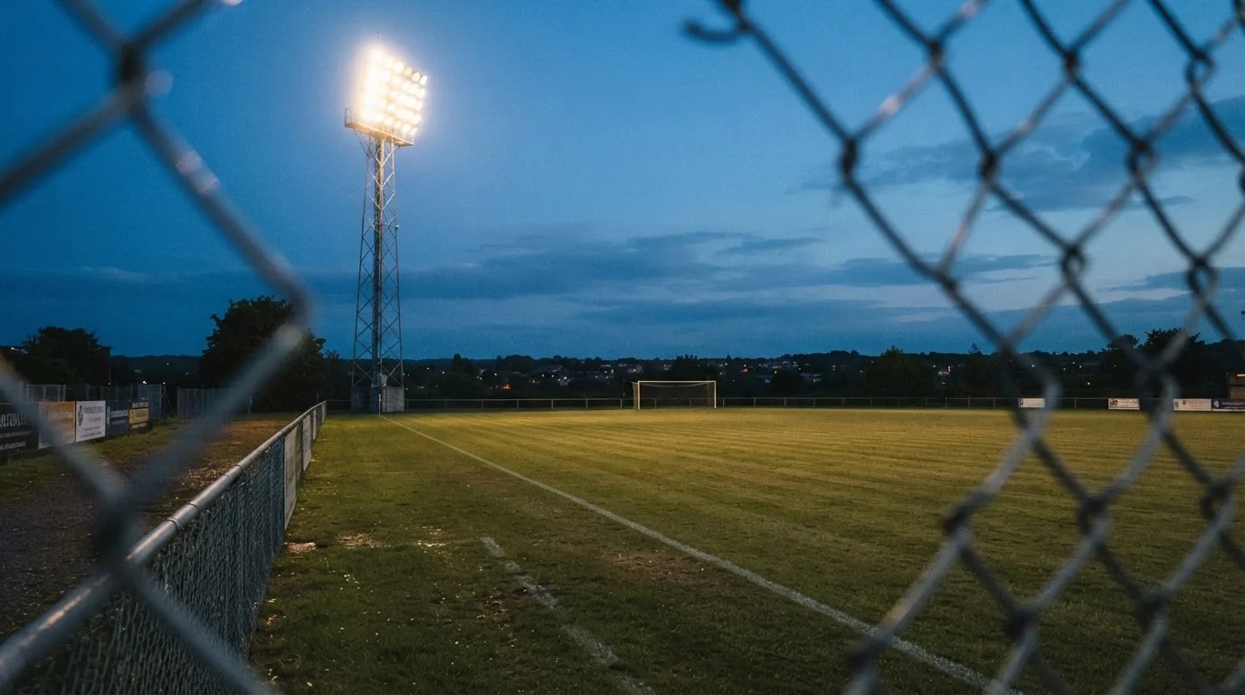 Eine Flutlichtanlage über einem leeren Sportplatz am Abend, im Vordergrund ein Maschendrahtzaun.