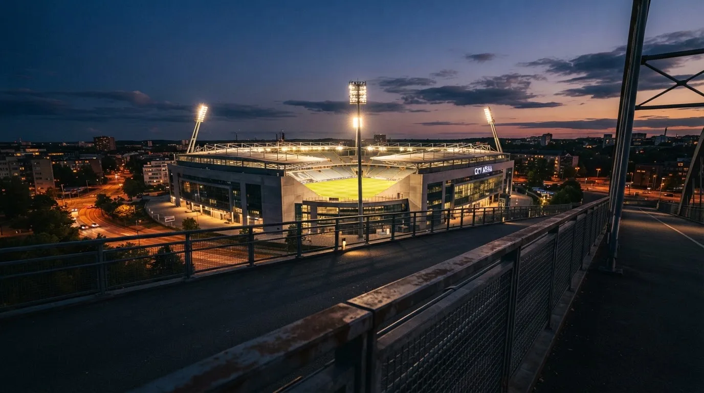 Ein Stadion bei Dämmerung mit eingeschalteten Flutlichtmasten, von einer Brücke aus aufgenommen.