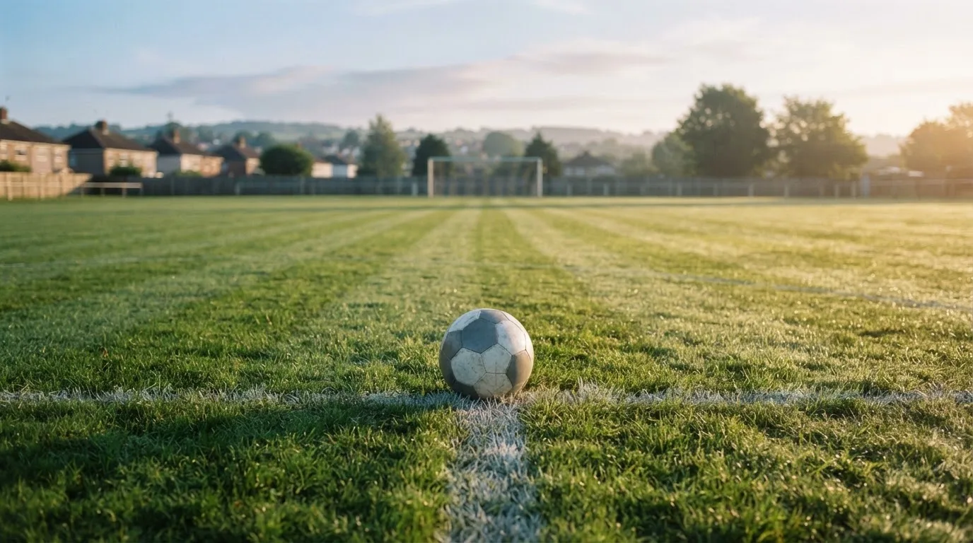 Ein Fußball auf dem Rasen eines leeren Sportplatzes am Rand einer Kleinstadt im Morgenlicht.