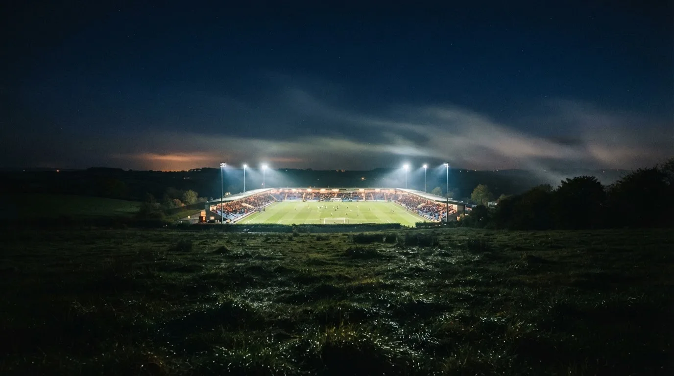 Ein Stadion unter Flutlicht, aus großer Entfernung über eine dunkle Wiese fotografiert, am Himmel zieht Nebel auf.