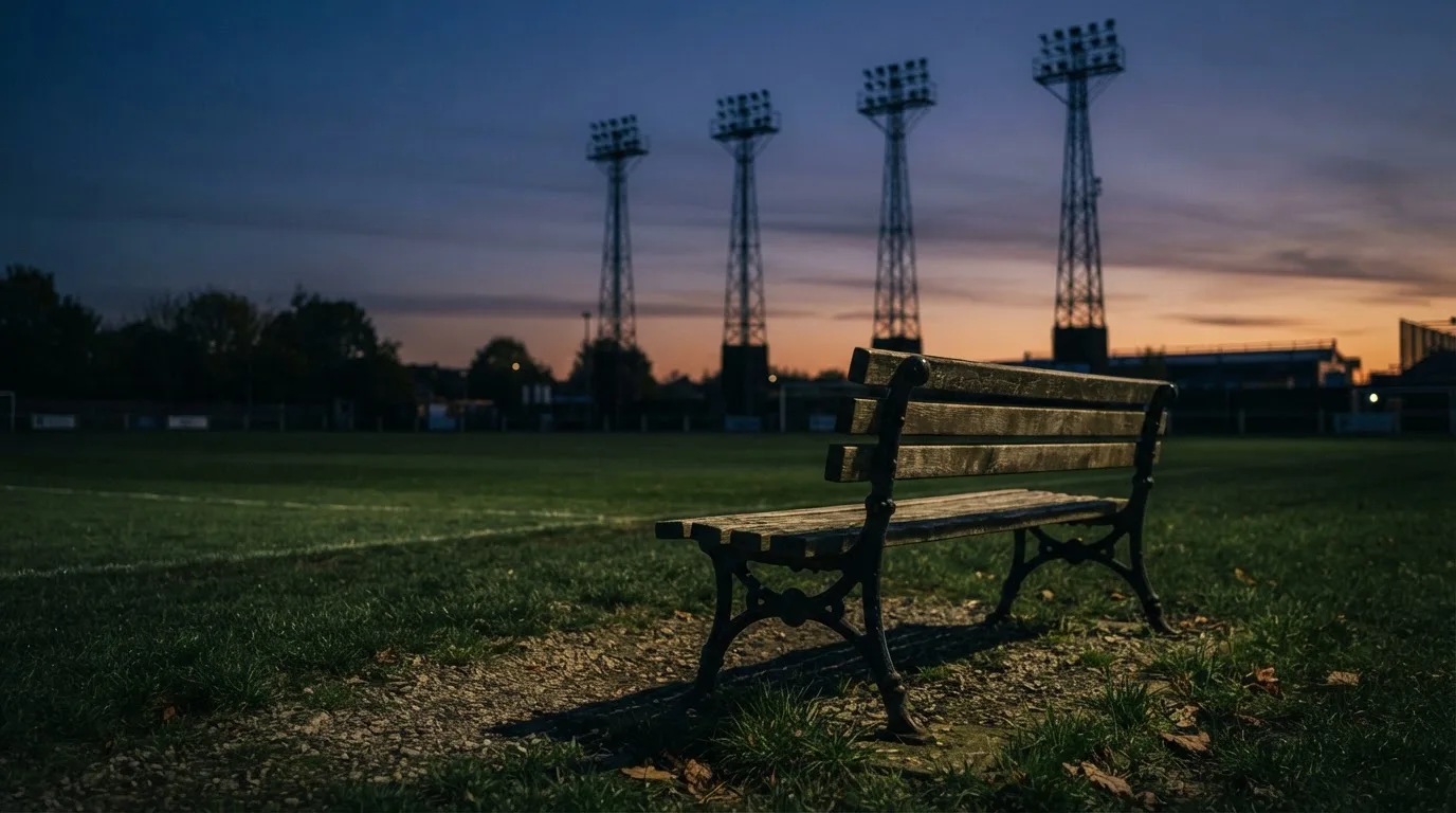 Eine leere Parkbank am Rand eines Fußballfelds in der Abenddämmerung, im Hintergrund ragen dunkle Flutlichtmasten in den Himmel.