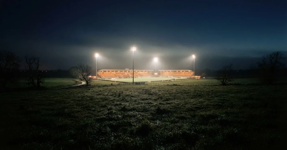 Ein Stadion unter Flutlicht, aus großer Entfernung über eine dunkle Wiese fotografiert, am Himmel zieht Nebel auf.