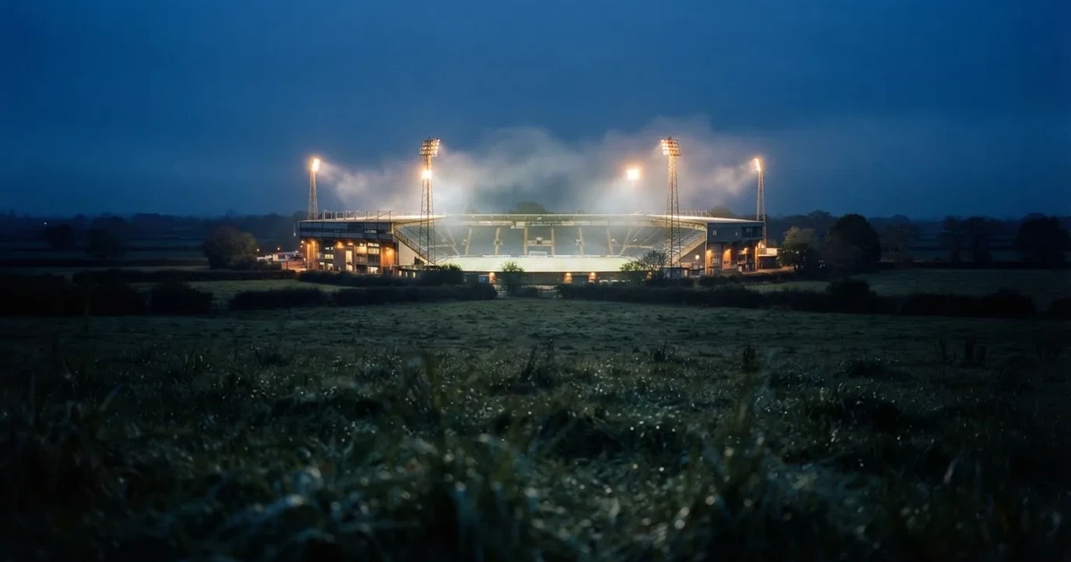 Ein Stadion unter Flutlicht, aus großer Entfernung über eine dunkle Wiese fotografiert, am Himmel zieht Nebel auf.