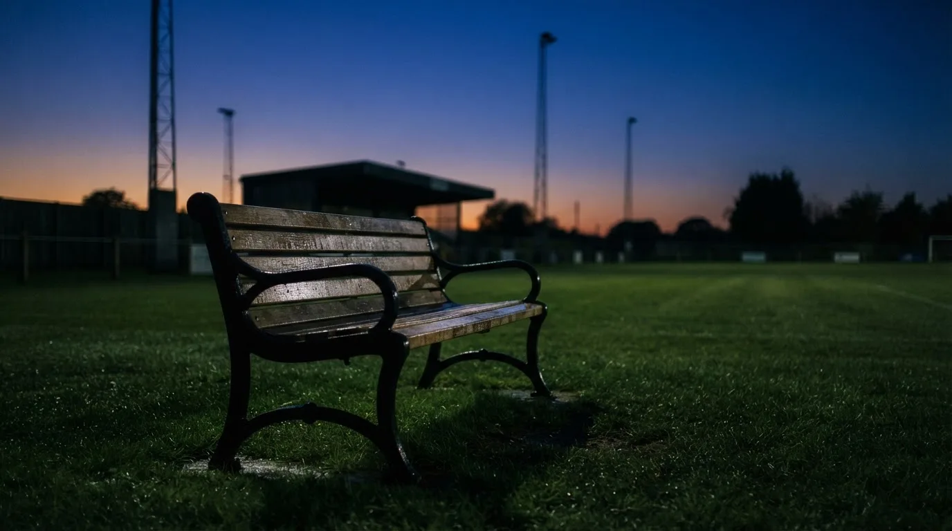 Eine leere Parkbank am Rand eines Fußballfelds in der Abenddämmerung, im Hintergrund ragen dunkle Flutlichtmasten in den Himmel.