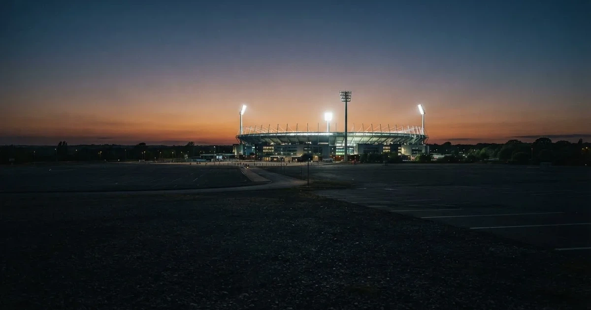 Ein Fußballstadion unter Flutlicht aus großer Entfernung, davor ein dunkler Parkplatz und ein heller Abendhimmel.