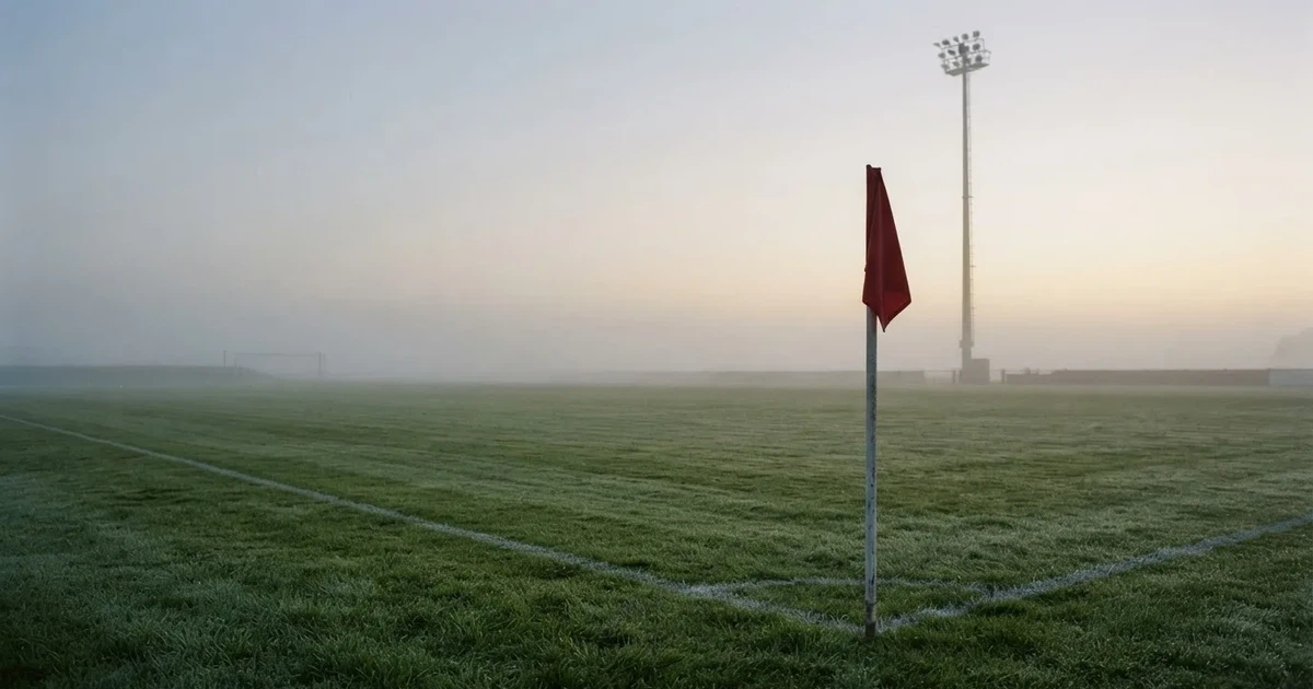 Eine Eckfahne steht im Nebel am Rand eines leeren Rasenplatzes in der Morgendämmerung, dahinter ein Flutlichtmast.