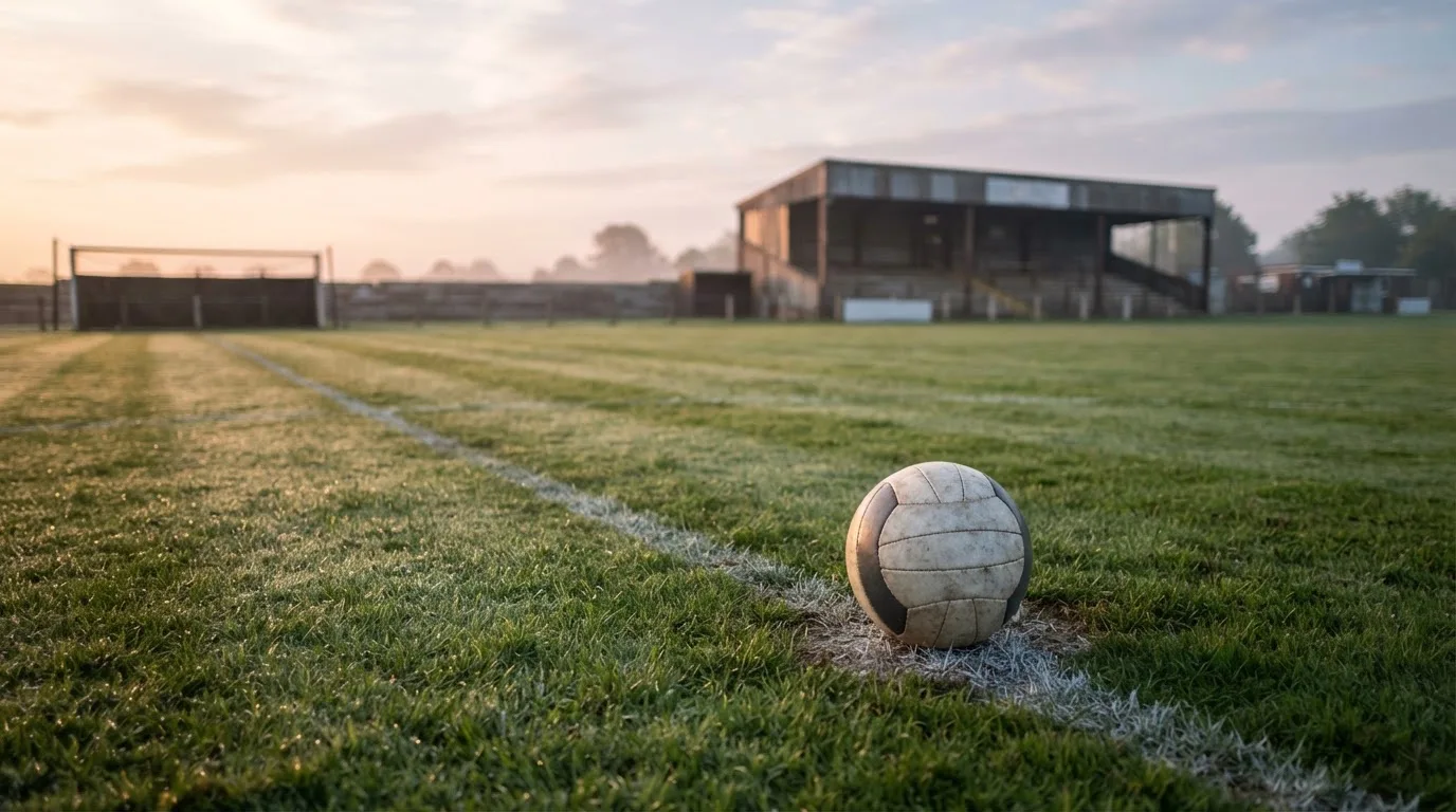 Ein Fußball liegt auf dem Anstoßpunkt eines leeren Rasenplatzes in der Morgendämmerung, im Hintergrund eine leere Tribüne.