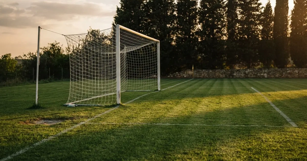 Ein Fußballtor mit Netz auf einem leeren Platz im Abendlicht, der Rasen frisch gemäht, im Hintergrund Zypressen.