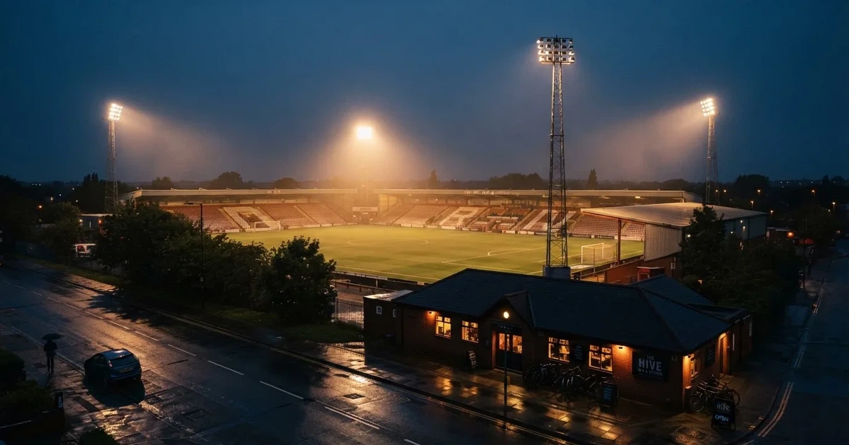 Ein Fußballstadion bei Flutlicht aus der Ferne gesehen, davor eine Straße und leuchtende Fenster eines Nebengebäudes.