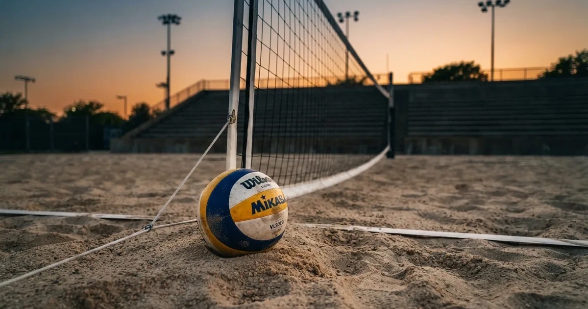 Ein Volleyball liegt auf dem Sand eines Beachvolleyball-Feldes neben dem gespannten Netz, im Hintergrund eine leere Tribüne bei Abendlicht.