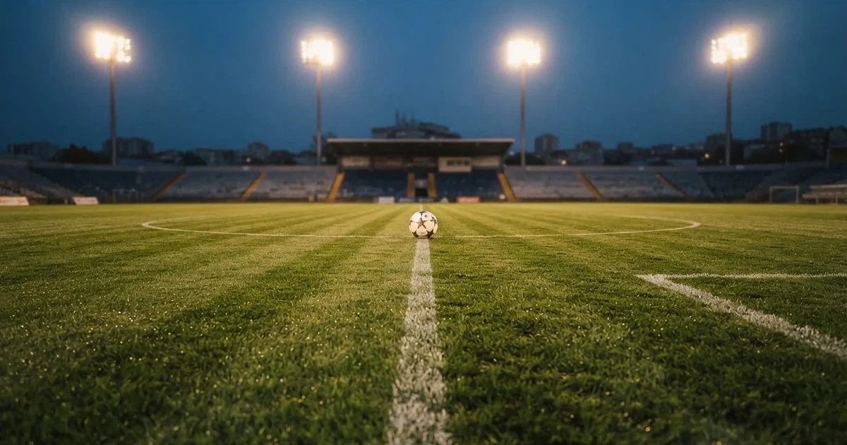 Ein Rasenplatz in der Dämmerung mit eingeschalteten Flutlichtmasten, ein Ball liegt am Anstoßpunkt, die Tribünen im Hintergrund sind unscharf.