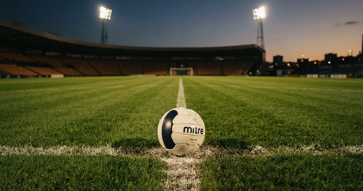 Ein Fußball liegt auf dem Anstoßpunkt eines leeren Rasenplatzes, im Hintergrund eine Tribüne und Flutlichtmasten in der Abenddämmerung.
