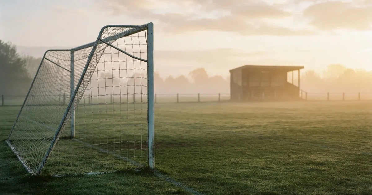 Ein Fußballtor mit Netz steht im Morgennebel auf einem leeren Rasenplatz, im Hintergrund die Silhouette einer kleinen Tribüne.