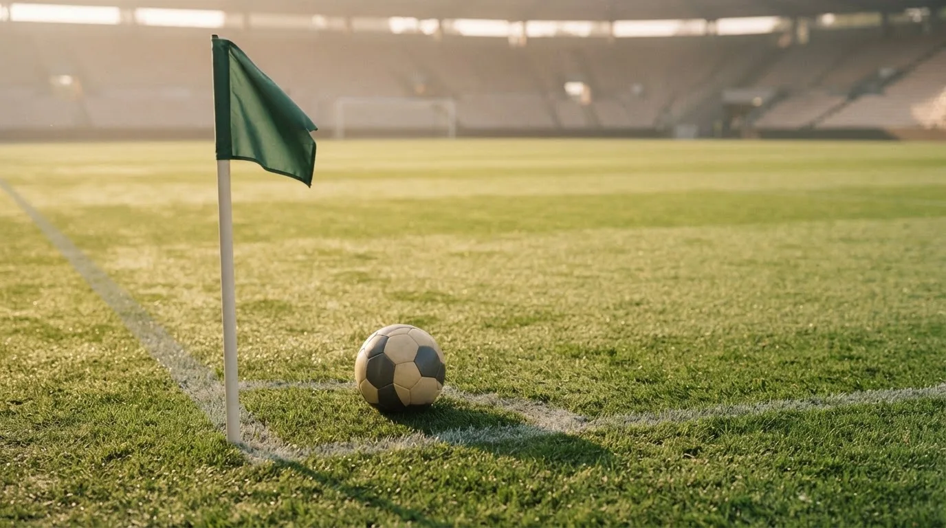 Ein Fußball liegt auf Rasen neben einer Eckfahne ohne Aufdruck auf einem leeren Platz im Morgenlicht.