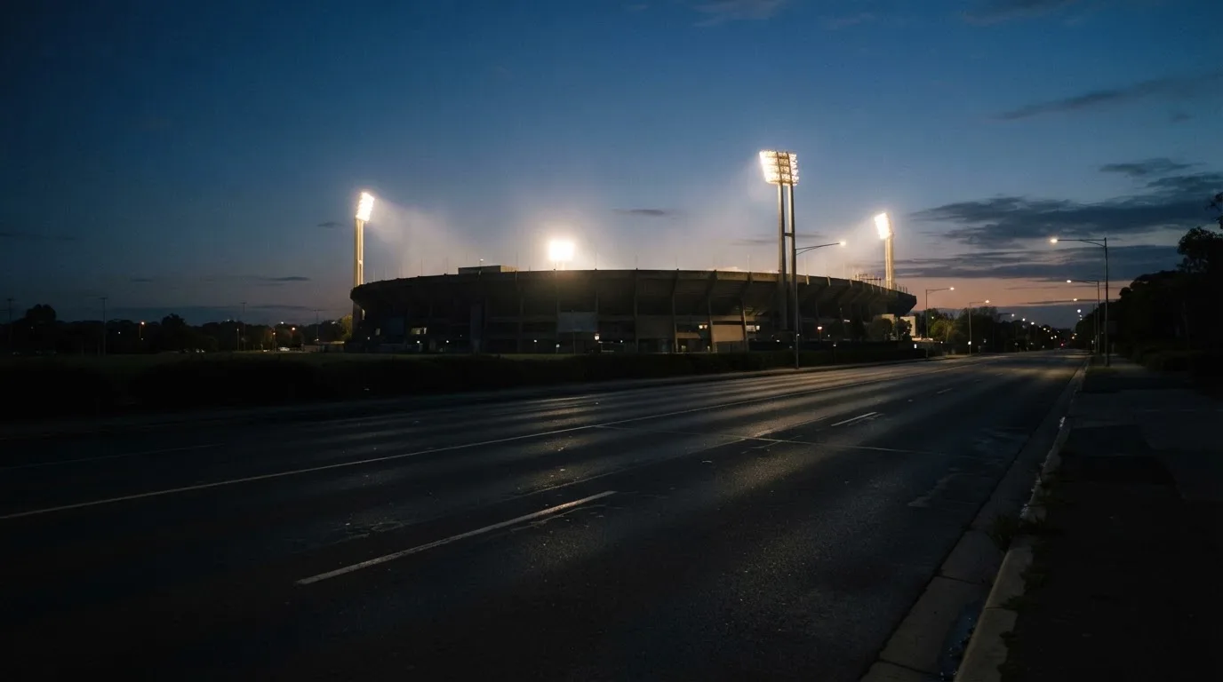 Ein Stadion unter Flutlicht aus großer Entfernung, davor eine leere Straße in der Abenddämmerung.