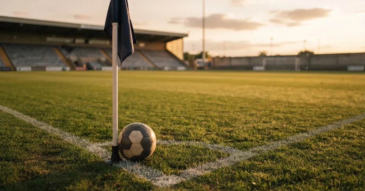 Ein Fußball liegt am Viertelkreis an der Eckfahne eines Rasenplatzes, im Hintergrund leere Tribünen in warmem Abendlicht.