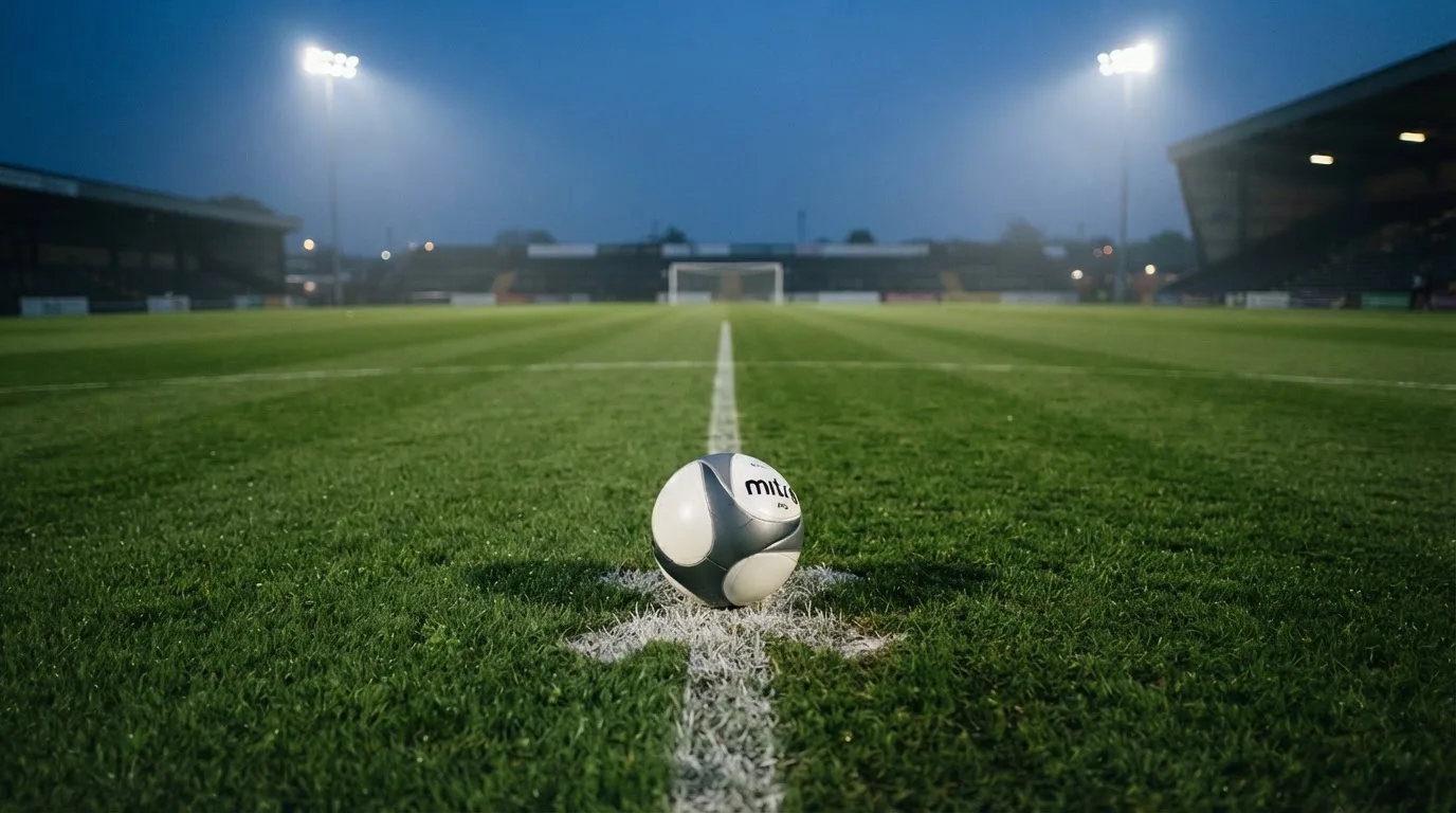 Ein Fußball liegt auf dem Mittelkreis eines leeren Rasenplatzes unter Flutlicht am frühen Abend, die Tribünen sind nur schemenhaft zu erkennen.