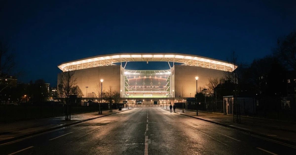 Ein hell erleuchtetes Fußballstadion bei Nacht aus der Ferne gesehen, über dem Dach ein klarer Himmel, davor eine leere Straße.