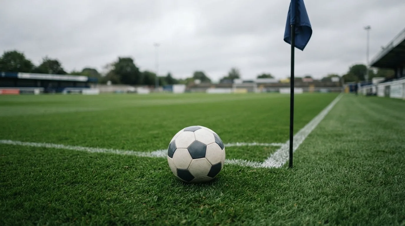 Ein Fußball liegt neben einer Eckfahne auf kurz gemähtem Rasen unter bewölktem Himmel.